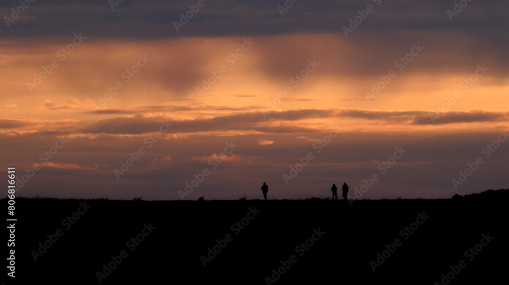 Silhouettes of three people, walking on top of a hill, golden hour sunset in the background and beautifully colored sky Silhouettes of three people, walking on top of a hill, golden hour sunset in the background and beautifully colored sky