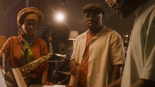 Medium tilting close-up of two African American male bandmates standing at keyboards in studio, trying out chords for new song, exchanging ideas, woman with saxophone watching and listening photo