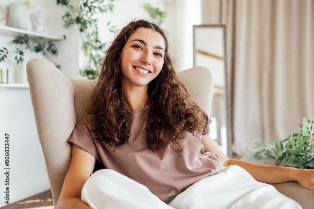 A charming curly-haired young woman in casual clothes is sitting in an armchair in the living room. An attractive brunette is smiling and relaxing at home. A charming curly-haired young woman in casual clothes is sitting in an armchair in the living room. An attractive brunette is smiling and relaxing at home.