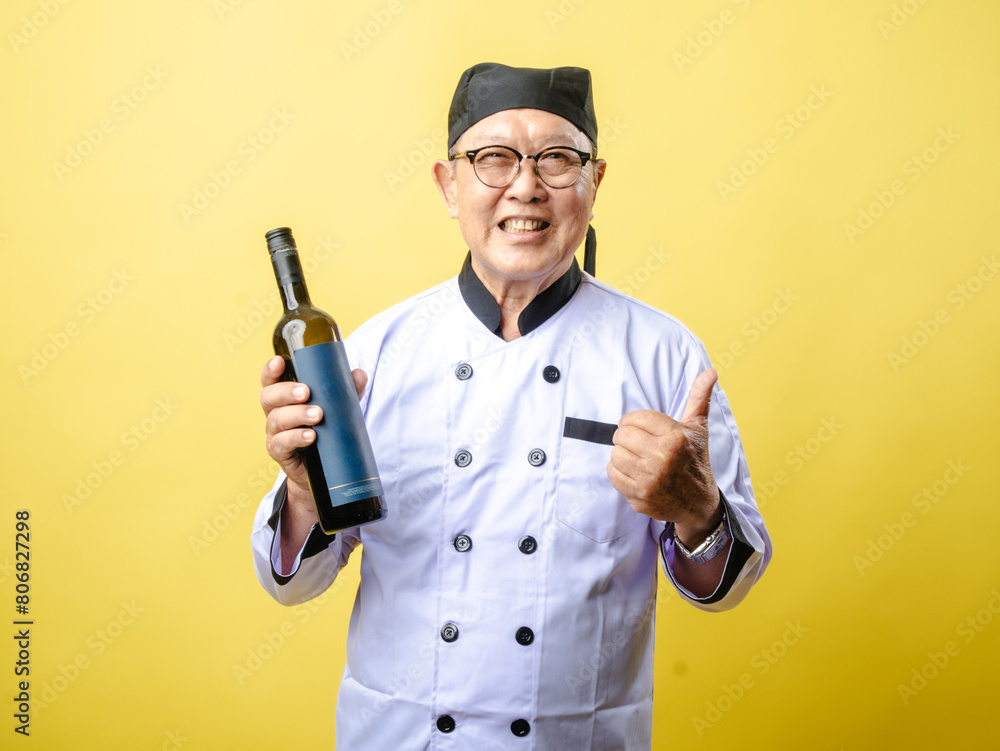 An elderly Asian chef in a white uniform and black hat smiles while holding a bottle of wine and giving a thumbs-up against a bright yellow background An elderly Asian chef in a white uniform and black hat smiles while holding a bottle of wine and giving a thumbs-up against a bright yellow background