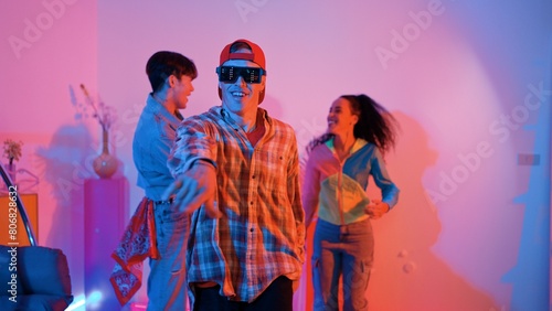Diverse happy people moving to lively music with neon light at studio. Attractive hipster dancing while asian man show energetic movement while multicultural team cheering and supporting. Regalement. photo