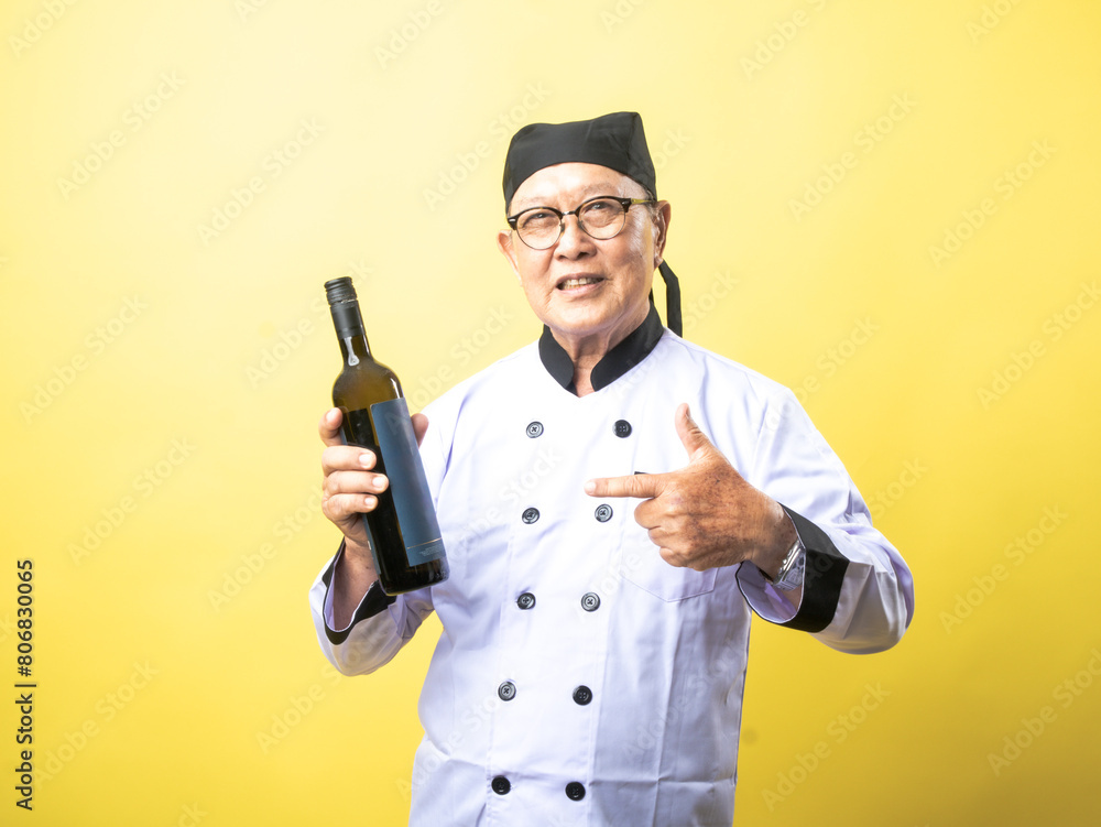 An elderly Asian chef in a white uniform and black hat smiles while holding a bottle of wine and giving a thumbs-up against a bright yellow background An elderly Asian chef in a white uniform and black hat smiles while holding a bottle of wine and giving a thumbs-up against a bright yellow background