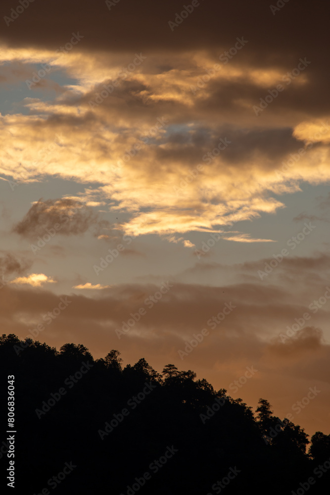 Landscape of thick clouds, orange light hitting the blue sky, silhouettes of trees, forests, and mountains. Area for text in the middle of the image