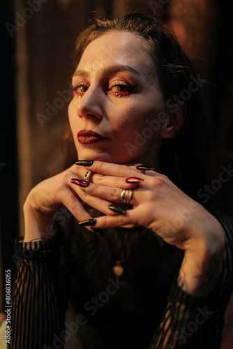 Closeup face of fortune teller with black nails sitting at table in dark esoteric room with mystical atmosphere, looking at camera with serious expression. Concept of prophecy, prediction and magic.