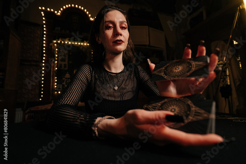 Low-angle portrait of fortuneteller shuffling deck of tarot cards by light of burning candles, looking at camera with confident expression. Concept of esoteric, fortune telling and predictions.