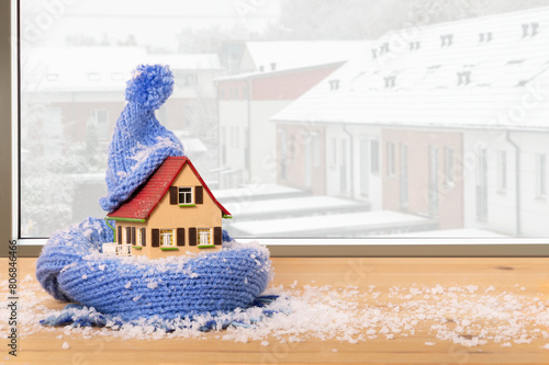snow-covered model house with hat and scarf on a window sill in front of a row of snow-covered houses in winter, thermal insulation of buildings