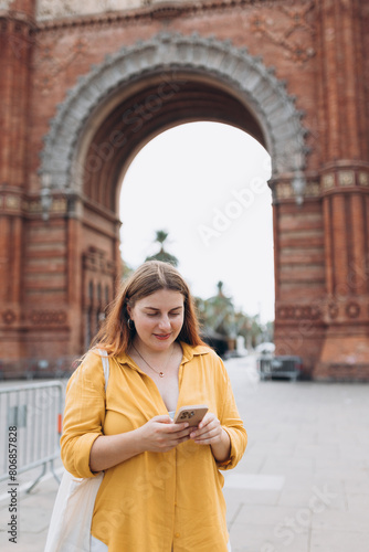 Woman with smart phone in summer vacation travel, standing near Barcelona triumphal arch. Female traveler visiting local landmarks. Concept of travel, tourism and vacation in city
