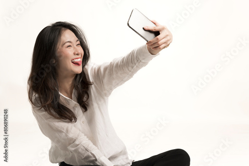 Portrait excited young woman taking a selfie in a studio isolated on white background.Beautiful asian woman laughing and showing stick out her tongue while taking selfie photo on cellphone.Asian cute.