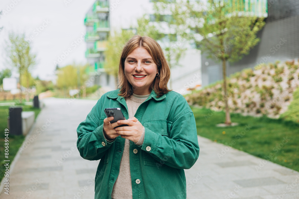30s cheerful woman tourist dressed in casual look holding smartphone gadget in hand for communicate on the city street. Happy woman chatting on phone and relaxing outdoors. Urban, people concept. 30s cheerful woman tourist dressed in casual look holding smartphone gadget in hand for communicate on the city street. Happy woman chatting on phone and relaxing outdoors. Urban, people concept.