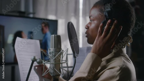 Medium side close-up of African American female voice-over actress in headphones reading dialog lines from cue sheet into microphone, while doing movie dubbing work in recording studio