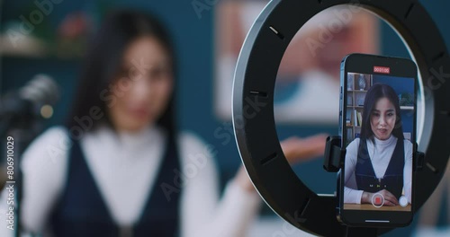 Camera focused of smartphone held by stand with circular lamp. Video recording. Young woman telling something in microphone or filming advertisement on phone. Showing gesture of love with hand. photo