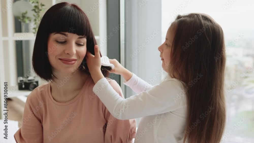 Little cute girl in casual outfit brushing her beautiful mothers hair sitting near big panoramic window in bedroom. Happy Caucasian family having fun on bad on weekend morning. Little cute girl in casual outfit brushing her beautiful mothers hair sitting near big panoramic window in bedroom. Happy Caucasian family having fun on bad on weekend morning.