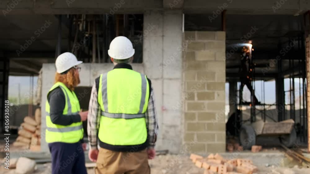 Two workers in vest and helmets chatting during a break at a construction site while standing against the backdrop of an unfinished house and welding work, copyspace.