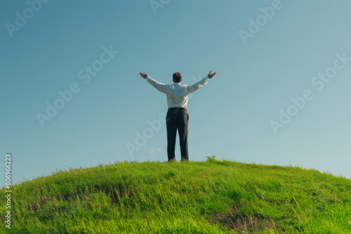 Businessman stands with arms outstretched on a grassy hilltop against a clear blue sky, symbolizing success, ambition, and the achievement of personal or career goals