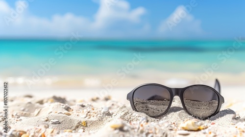  A pair of sunglasses lies on the sandy beach, surrounded by the ocean in the foreground and a clear, blue sky overhead