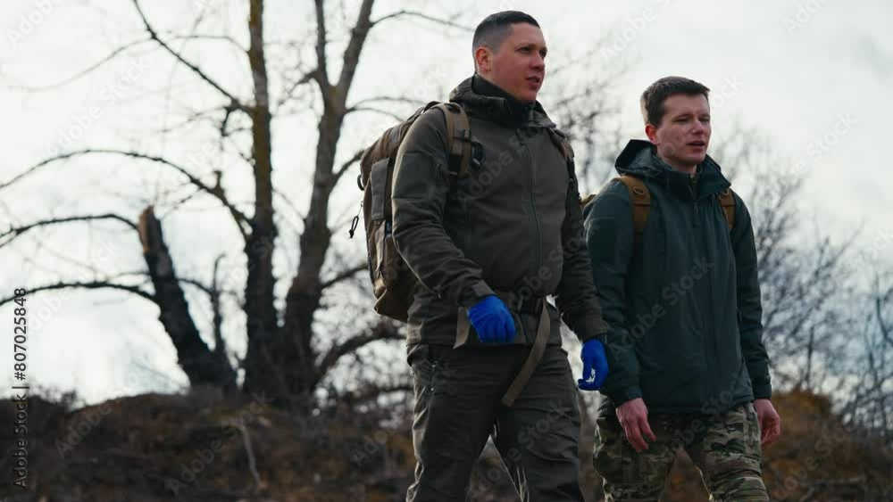 Side view of two confident young men in dark green army uniforms and blue medical gloves walking along the military training ground and combat practice and communicating with backpacks on their backs Side view of two confident young men in dark green army uniforms and blue medical gloves walking along the military training ground and combat practice and communicating with backpacks on their backs