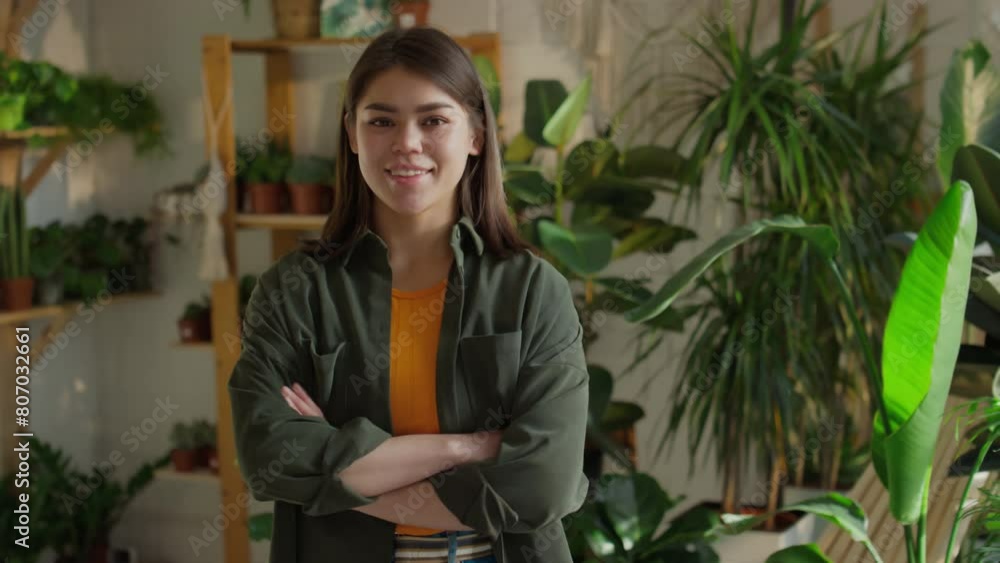 Portrait of smiling Caucasian sales assistant or entrepreneur posing with crossed arms in front of stock of green potted plants displayed on wall shelves Portrait of smiling Caucasian sales assistant or entrepreneur posing with crossed arms in front of stock of green potted plants displayed on wall shelves