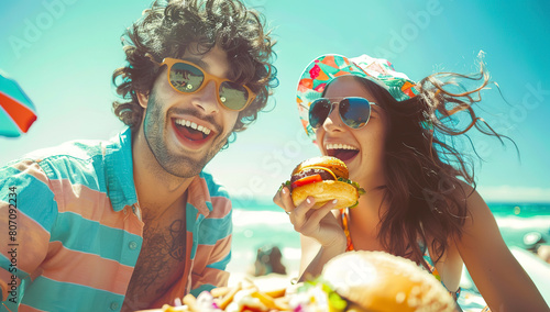Happy friends are having lunch at the beach, enjoying a burger and beer on a table with summer food in the background. It is a vacation concept
