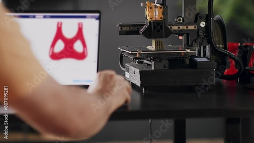 A man graphic designer develops a model of postoperative fixation bandage in red colour for the heart and chest for women, depicted on a laptop screen. A 3d printer machine works in automated photo