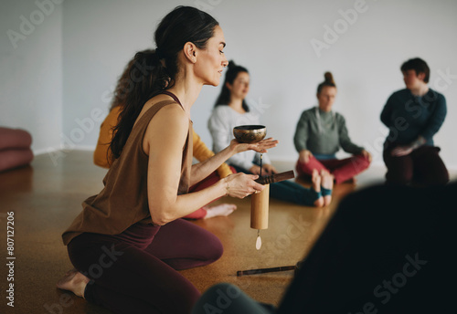 Female yoga instructor using a singing bowl and chime during a meditation class of students sitting together on the floor of her studio photo