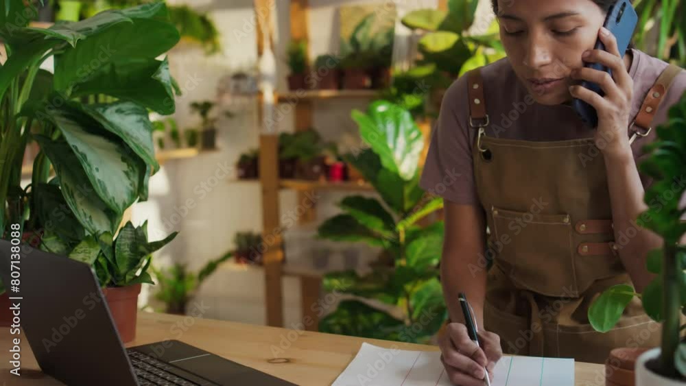 Medium tilt of Hispanic young woman wearing apron working as plant shop salesperson taking order from customer, typing on laptop and making notes during phone talk, leafy atmosphere in background Medium tilt of Hispanic young woman wearing apron working as plant shop salesperson taking order from customer, typing on laptop and making notes during phone talk, leafy atmosphere in background