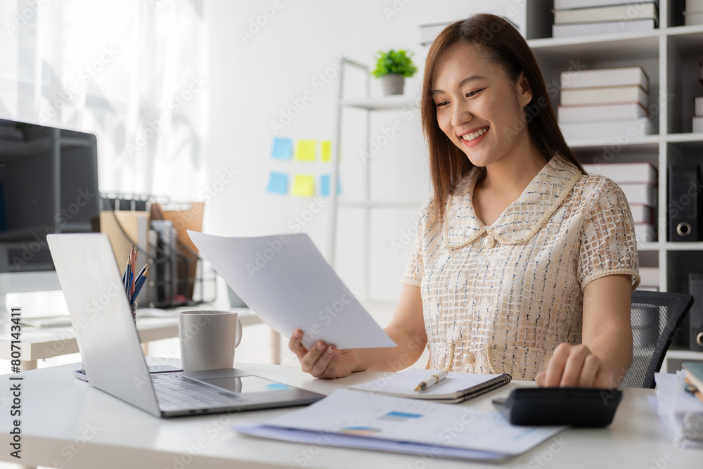 Beautiful Asian female accountant smiling while working with laptop and documents about finance, mathematics, accounting reports, statistics. and research ideas Beautiful Asian female accountant smiling while working with laptop and documents about finance, mathematics, accounting reports, statistics. and research ideas