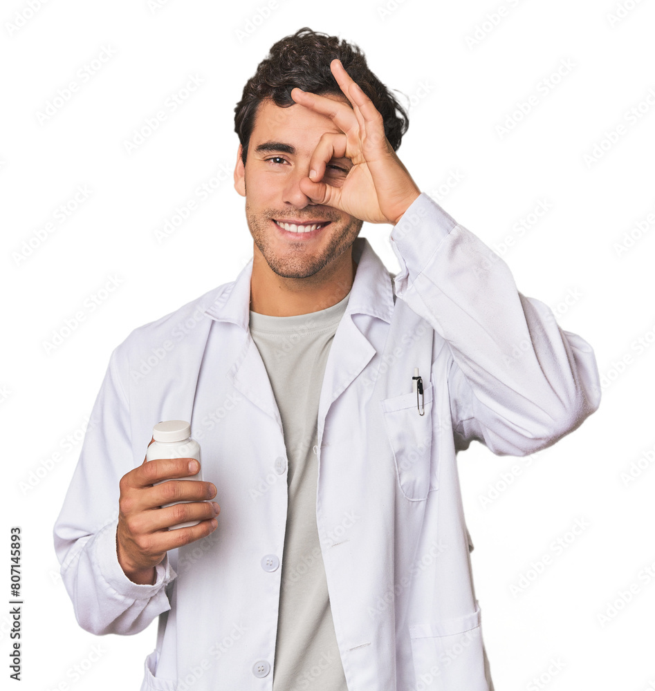 Young Hispanic pharmacist with pills excited keeping ok gesture on eye. Young Hispanic pharmacist with pills excited keeping ok gesture on eye.