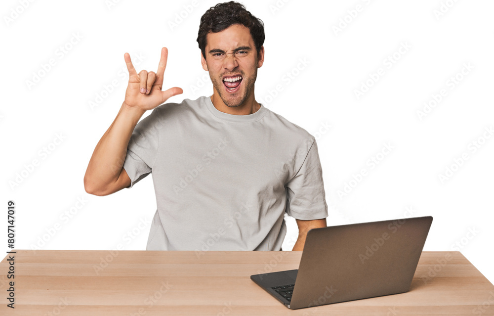 Hispanic young man working on laptop showing a horns gesture as a revolution concept. Hispanic young man working on laptop showing a horns gesture as a revolution concept.