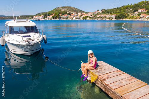 Cute little girl kid sits on wooden pier next moored boat yacht with scenic turquoise sea water background. Happy child in cap hold fishing net have fun explore nature at summer vacation