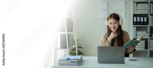 A woman is sitting at a desk with a laptop and a book. She is wearing headphones and she is focused on her work. The room is filled with bookshelves