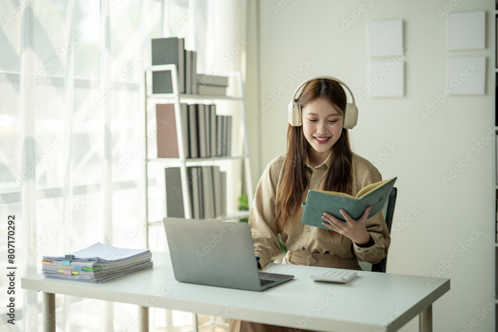 Fototapeta premium A woman is sitting at a desk with a laptop and a book. She is wearing headphones and smiling. The scene suggests that she is enjoying her work or leisure time