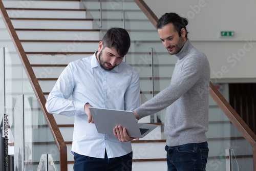 deux jeune hommes d'affaire ou employés de bureau discutent debout dans les locaux de leur entreprise. l'un d'eux porte un ordinateur portable photo