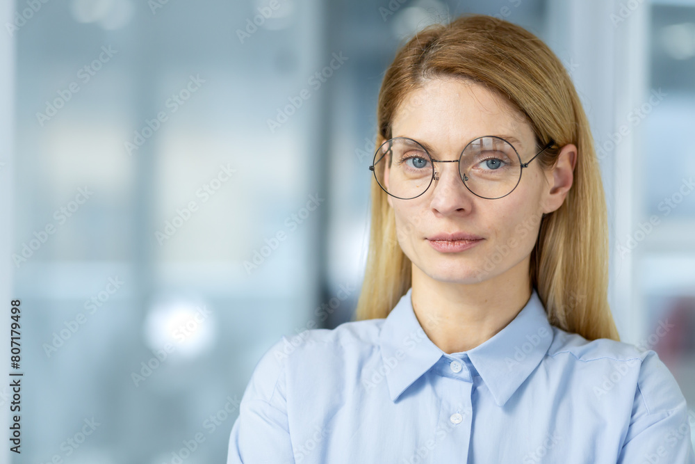 Confident professional businesswoman in a blue blouse and round glasses, standing in a modern office environment with a focused expression. Confident professional businesswoman in a blue blouse and round glasses, standing in a modern office environment with a focused expression.