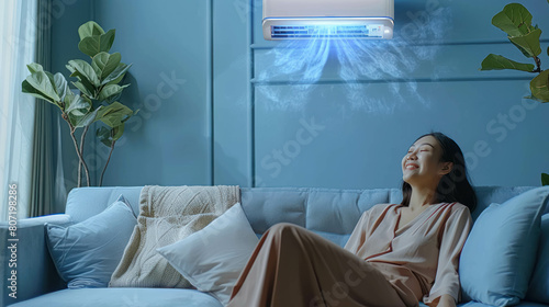 Smiling Asian Woman Lounges On A Sofa In A Living Room Enjoying The Comfort Of An Efficient Air Conditioner photo