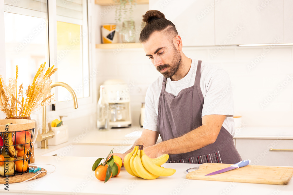 A man is cutting a banana in a kitchen. He is wearing an apron and has a knife in his hand. There are several fruits on the counter, including bananas, oranges, and apples A man is cutting a banana in a kitchen. He is wearing an apron and has a knife in his hand. There are several fruits on the counter, including bananas, oranges, and apples
