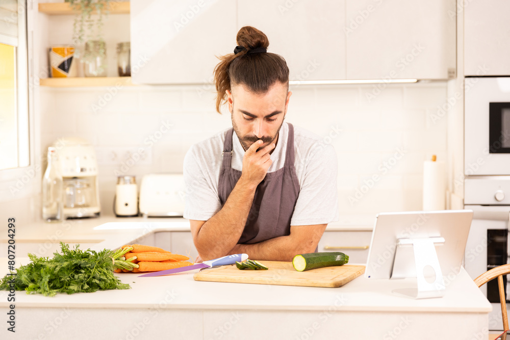A man is sitting at a kitchen counter with a tablet in front of him. He is looking at a carrot and a cucumber on the cutting board A man is sitting at a kitchen counter with a tablet in front of him. He is looking at a carrot and a cucumber on the cutting board