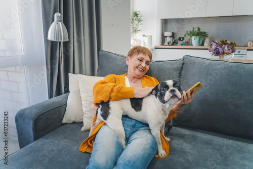 Elderly Caucasian woman hugging her pet French bulldog while sitting on the sofa at home, love for pets © olezzo