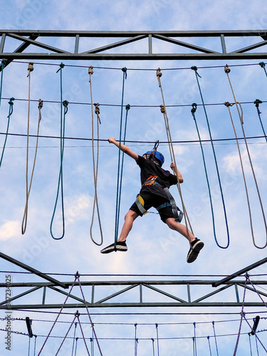 A teenage boy in sports safety equipment crosses a suspension rope bridge in a rope amusement park. View from below on the background of the sky
