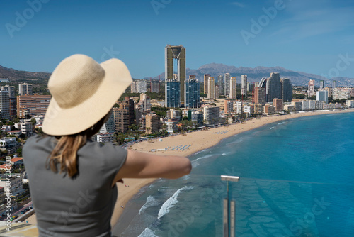 Woman standing at viewpoint called Mirador de l'Ermita Verge del Mar - looking at the panorama of Benidorm (Costa Blanca), West Beach Promenade (Playa de Poniente). Wearing a straw hat and khaki dress photo