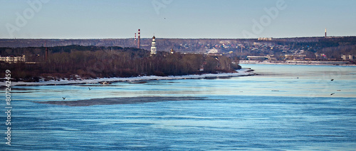 Winter city landscape. Panorama. On the nearest bank of a wide river, in a grove there is an Orthodox temple, buildings and industrial pipes. There is a settlement on the opposite bank.