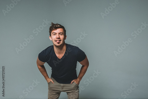 A man on a gray background in a dark blue T-shirt, hands in pockets, leans forward and shows his tongue. A man is messing around on camera photo