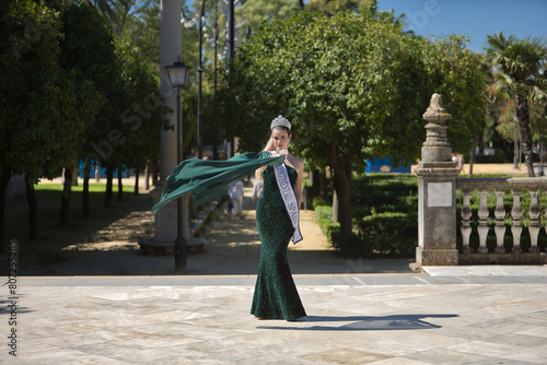Young, pretty, blonde woman in a green party outfit with sequins, a diamond crown and a beauty pageant winner's sash, twirling so that the air blows her cape. Concept of beauty, fashion, pageant.
