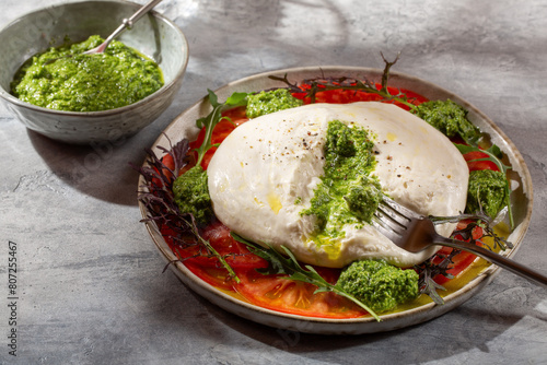 Tomato salad with burrata cheese and green pesto on gray background.