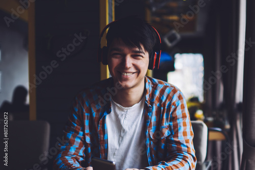 Happy male meloman listening favourite music in stylish modern headphones while holding smartphone in hands sitting at wooden table with cup of tasty coffee and books in coworking space photo