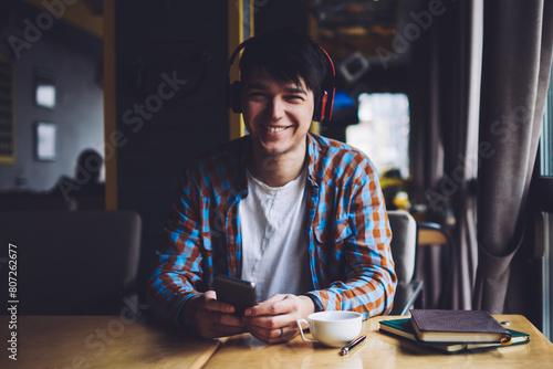 Happy male meloman listening favourite music in stylish modern headphones while holding smartphone in hands sitting at wooden table with cup of tasty coffee and books in coworking space photo