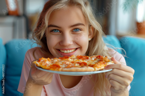 Close up view of happy hungry pretty blonde hair girl sitting on blue sofa at home and eating tasty pizza 