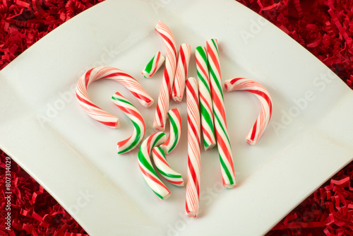 Red and white and green broken candy canes from poor packaging on white snack plate on background of red crinkled shredded paper