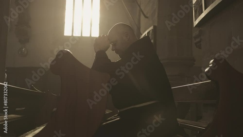 Monk in robe praying on pew in church side view medium shot. Christian clergyman with closed eyes and clasped hands sitting on wooden bench in monastery photo