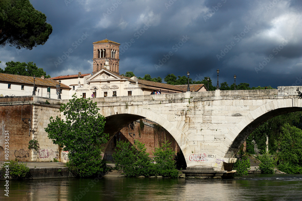 Ponte Fabricio in Rome which connects with the Tiber Island, bell tower of the Basilica of San Bartolomeo in the background, dark sky full of clouds. Ponte Fabricio in Rome which connects with the Tiber Island, bell tower of the Basilica of San Bartolomeo in the background, dark sky full of clouds.