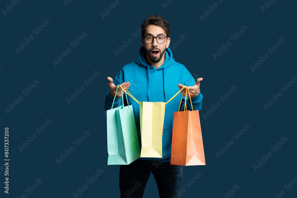 A young man with a beard and glasses expresses surprise and excitement as he holds several colorful shopping bags. He is dressed in a casual blue hoodie and stands against a solid dark blue background A young man with a beard and glasses expresses surprise and excitement as he holds several colorful shopping bags. He is dressed in a casual blue hoodie and stands against a solid dark blue background